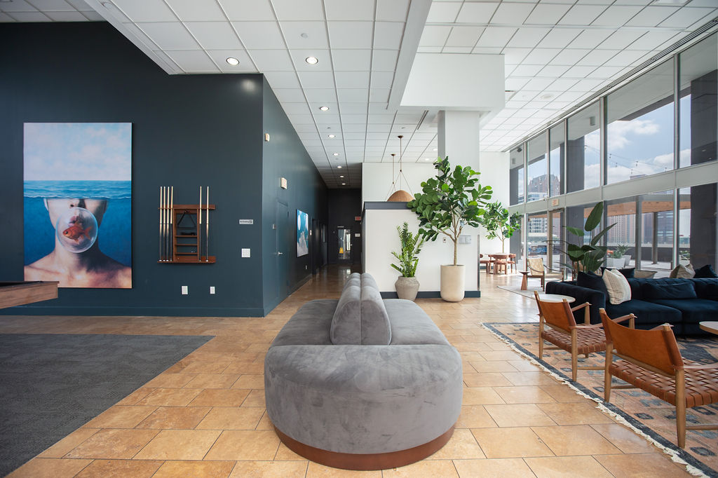 Modern lobby with large windows, gray curved sofa, lounge chairs, indoor plants, wall art of a person blowing bubblegum, billiard cues on the wall, and tiled floor with natural light filling the space—an inviting glimpse into upscale apartments in Richmond TX.